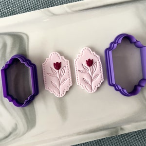 May include: Two pink fondant cookie cutters with a tulip design and a purple plastic cookie cutter in the shape of a rectangle.