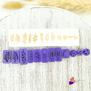 Peut inclure: Une collection de tampons violets et dorés en forme de fleurs et de feuilles. Les tampons sont disposés sur une surface blanche. Les tampons dorés sont sur une bande blanche, tandis que les tampons violets sont des blocs individuels. Les motifs comprennent diverses feuilles, fleurs et une libellule.