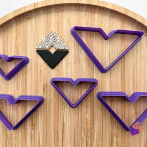 Peut inclure: Cinq emporte-pièces en plastique violet en forme de cœur avec un fond pointu. Les emporte-pièces sont disposés sur une surface en bois.