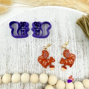 May include: A pair of squirrel-shaped earrings and two purple cookie cutters. The earrings are brown with gold-colored hardware and feature a squirrel design with floral accents and an acorn. The cookie cutters are purple and also shaped like squirrels.