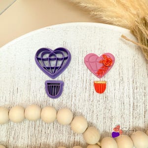 May include: Two heart-shaped cookie cutters, one purple and one pink, with a basket design. The cutters are for making heart-shaped hot air balloons.