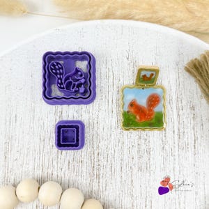May include: Three items are displayed: a purple squirrel-shaped cookie cutter, a small square purple stamp, and a squirrel-themed pendant. The pendant features a painted squirrel on a green field with a blue sky, framed in gold.