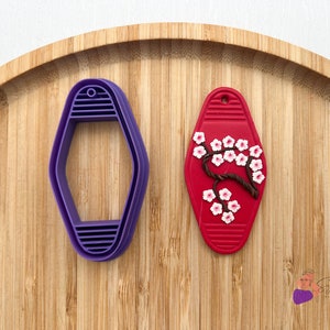 May include: A purple plastic hexagonal cookie cutter and a red silicone mold with a white cherry blossom design. Both are on a wooden surface.