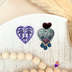 Pode incluir: Um cortador de biscoitos roxo em forma de coração e um brinco em forma de coração. O cortador de biscoitos tem um design de rosto triste. O brinco tem um coração listrado em preto e branco com um rosto triste, lágrimas e um coração vermelho escuro no topo.