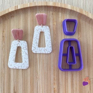 Puede incluir: Un par de pendientes de arcilla polimérica blanca y rosa con un patrón de hojas y un cortador de plástico morado con la forma de los pendientes.