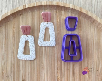 Op de afbeelding: Een paar witte en roze polymer clay oorbellen met een bladmotief en een paarse plastic uitsteker in de vorm van de oorbellen.