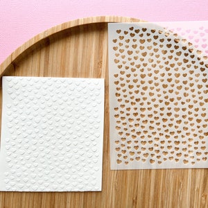 Peut inclure: Deux feuilles de papier blanches avec un motif de cœurs. Une feuille a des cœurs en relief et l'autre est un pochoir avec des cœurs découpés.