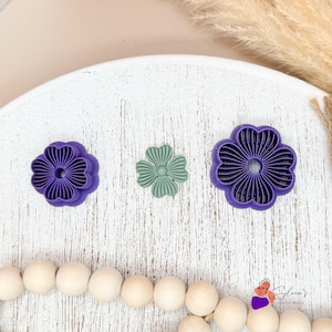 Peut inclure: Trois emportes-pièces en forme de fleur, deux sont violets et un est vert. Les emportes-pièces sont sur une surface en bois blanc.