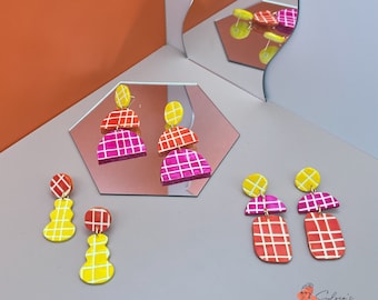 May include: Three pairs of colorful geometric polymer clay earrings. The earrings are in shades of yellow, red, and pink. The earrings are all different shapes and sizes. The earrings are displayed on a light pink surface.