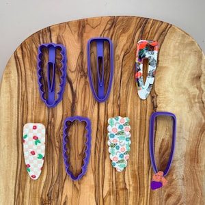 Peut inclure: Des emporte-pièces en plastique violet pour pinces à cheveux sur une surface en bois. Les emporte-pièces sont de formes et de tailles différentes. Il y a aussi plusieurs pinces à cheveux à motifs.