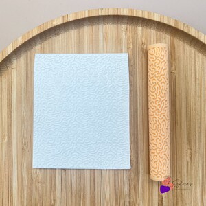 Op de afbeelding: Een lichtblauwe, textuurrijke siliconenmat en een oranje siliconen deegroller met een textuurontwerp. Beide items bevinden zich op een houten oppervlak.