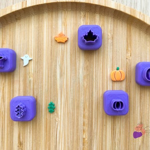 Puede incluir: Cortadores de galletas de plástico morado en varias formas, incluyendo un fantasma, una calabaza, una hoja y un árbol. Los cortadores están dispuestos sobre una superficie de madera.