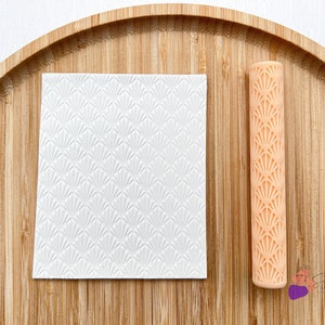 May include: A white textured square with a fan-shaped pattern and a peach-colored rolling pin with the same pattern. The rolling pin and square are on a wooden surface.