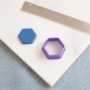 Puede incluir: Dos cortadores de galletas en forma de hexágono, uno azul y otro morado, sobre una superficie de baldosas blancas.