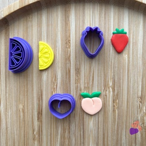 Peut inclure: Un ensemble de six emporte-pièces en plastique violet et jaune en forme de citron, de fraise, de pêche et de cœur. Les emporte-pièces sont disposés sur une surface en bois.
