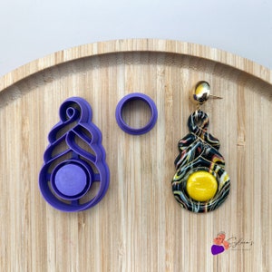 Op de afbeelding: Een polymeerklei-oorbel en bijpassende paarse kleisnijders liggen op een houten dienblad. De oorbel heeft een gele kern en een zwart, wit en rood gemarmerd ontwerp. De snijders zijn voor het maken van de oorbelvorm.