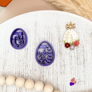 Könnte beinhalten: Drei Artikel zum Thema Ostern auf einer weißen Holzoberfläche. Zwei lila eiförmige Ausstecher mit floralen Designs und ein dekorierter eiförmiger Ohrring mit einer goldenen Schleife, goldenen Blättern und kleinen roten und rosa Blumen.