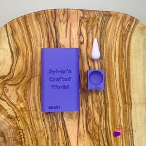 Può includere: Stampi in silicone viola per l'artigianato, con il testo "Sylvia's Crafted World" e il codice "BR001" sullo stampo più grande. Lo stampo più piccolo è rotondo e ha il codice "BR001" su di esso. È inclusa anche una forma a goccia di silicone bianco.