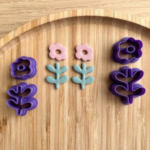 Peut inclure: Un ensemble d'emporte-pièces en forme de fleurs et de feuilles de couleur violette et rose sur une surface en bois. Les emporte-pièces sont en plastique et sont conçus pour découper des formes dans la pâte.