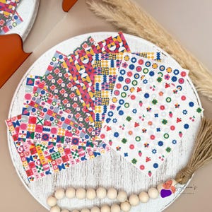 Puede incluir: Una colección de hojas de papel estampadas con varios diseños, incluyendo patrones florales, geométricos y abstractos. La paleta de colores incluye rojo, rosa, azul, amarillo y verde. Los papeles están dispuestos sobre una bandeja de madera blanca envejecida.