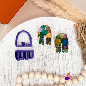 May include: A pair of colourful, abstract earrings with a rainbow design. The earrings feature a mix of green, blue, yellow, and orange hues. A purple cookie cutter is next to the earrings. The earrings are on a white wooden surface.