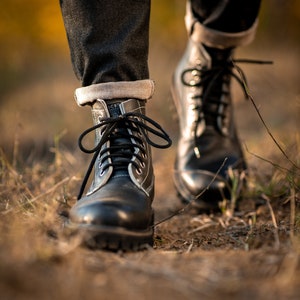 May include: Close-up of a pair of black leather boots with white stitching and black laces. The boots are worn by a person walking on a dirt path.