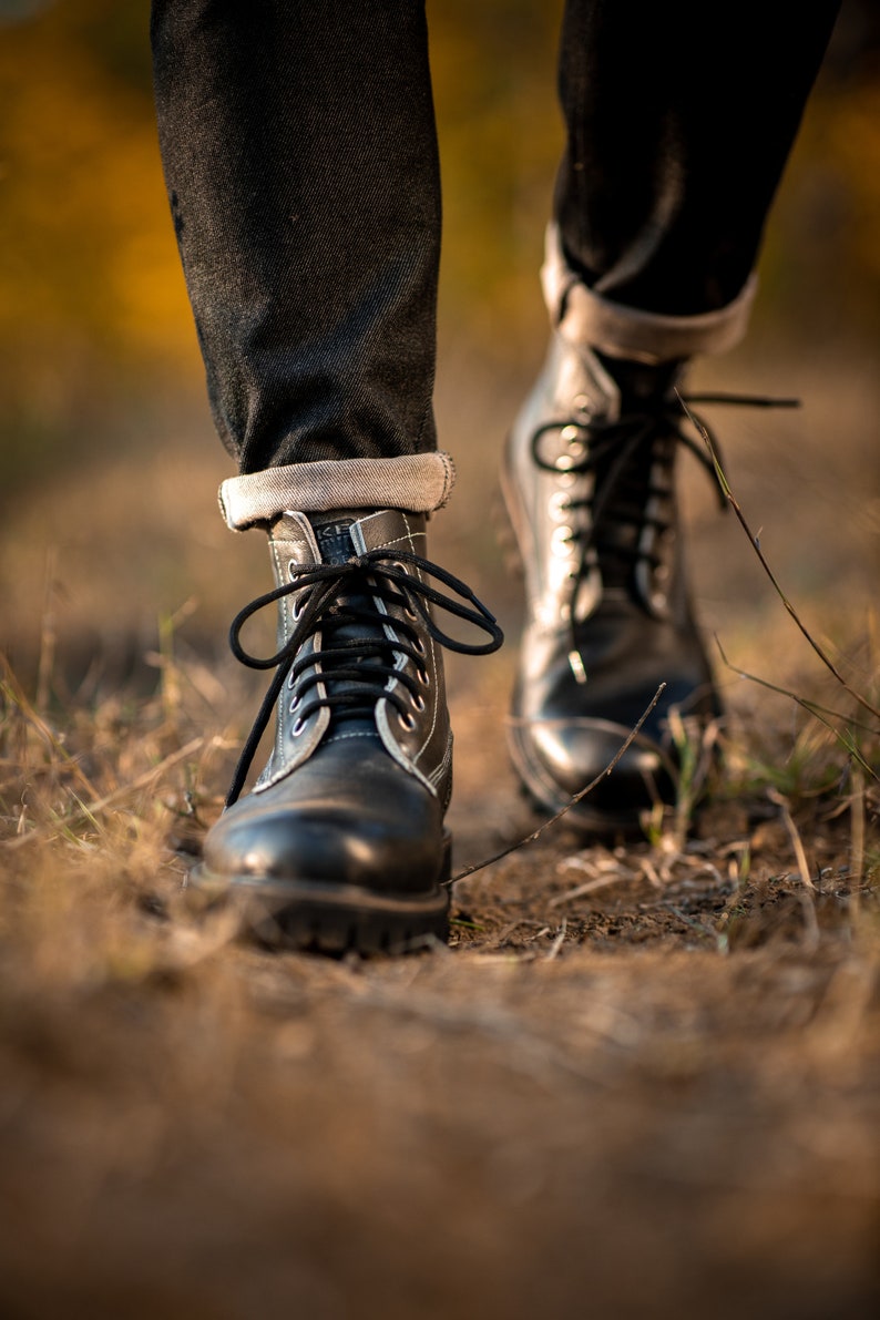May include: Close-up of a pair of black leather boots with white stitching and black laces. The boots are worn by a person walking on a dirt path.