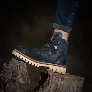 May include: A dark blue suede hiking boot with a brown rubber sole and a lace-up closure. The boot is resting on a weathered tree stump.