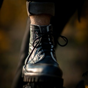 May include: Close-up of a black leather boot with white stitching and laces. The boot is worn and has a worn-in look. The boot is resting on a dark surface.