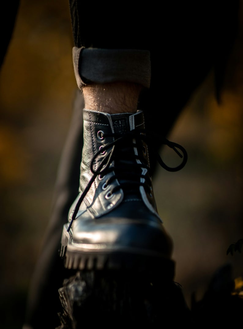 May include: Close-up of a black leather boot with white stitching and laces. The boot is worn and has a worn-in look. The boot is resting on a dark surface.