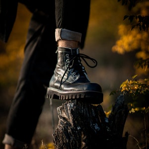May include: A black leather boot with white laces is perched on top of a weathered tree stump. The boot has a thick sole and a rounded toe. The background is blurred and out of focus, suggesting a natural setting.