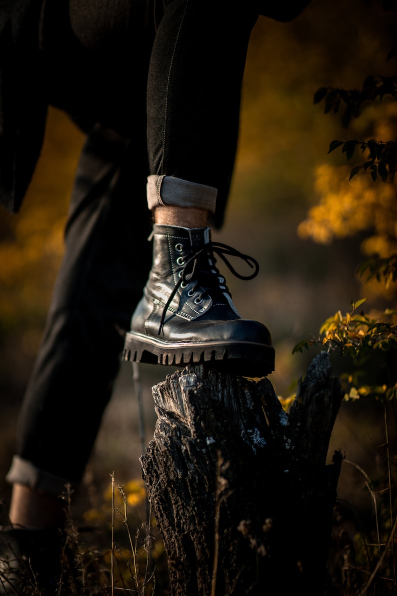 May include: A black leather boot with white laces is perched on top of a weathered tree stump. The boot has a thick sole and a rounded toe. The background is blurred and out of focus, suggesting a natural setting.