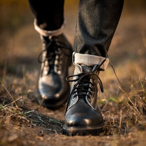 May include: Close-up of a pair of black leather boots with laces. The boots are worn by a person walking on a dirt path with dry grass.