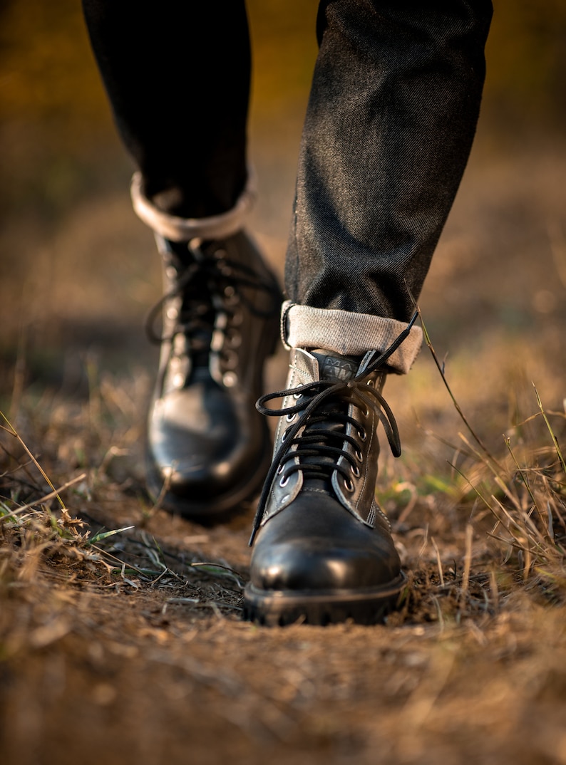 May include: Close-up of a pair of black leather boots with laces. The boots are worn by a person walking on a dirt path with dry grass.