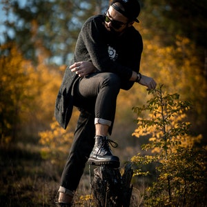 May include: A man wearing a black sweater, black jeans, and black boots stands on a tree stump in a forest. The background is blurred and shows trees with yellow leaves.