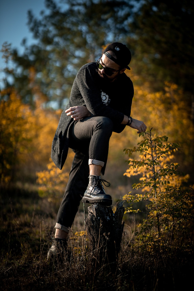 May include: A man wearing a black sweater, black jeans, and black boots stands on a tree stump in a forest. The background is blurred and shows trees with yellow leaves.