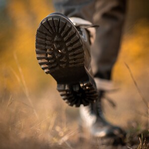 May include: Close-up of a black leather boot with a thick, rugged sole. The boot is being worn by a person walking on a path covered in dry grass.