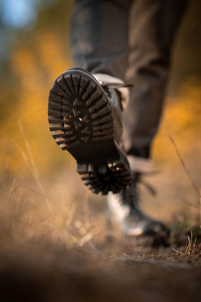 May include: Close-up of a black leather boot with a thick, rugged sole. The boot is being worn by a person walking on a path covered in dry grass.