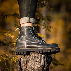 May include: Black leather lace-up boots with a thick sole, resting on a tree stump. The boots have a white label with the text "MANMADE" on the side.