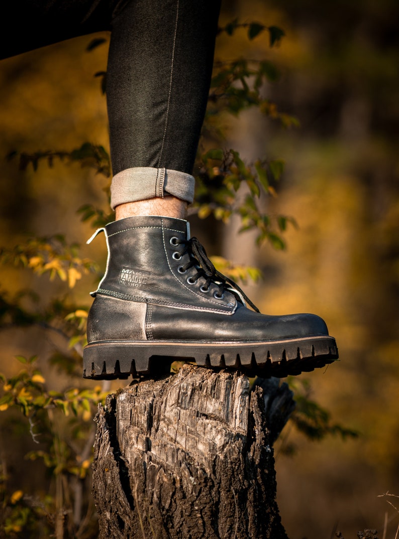 May include: Black leather lace-up boots with a thick sole, resting on a tree stump. The boots have a white label with the text "MANMADE" on the side.