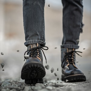 May include: Close-up of a person wearing black leather boots with brown laces and dark blue jeans. The boots are on a rocky surface with small stones scattered around.