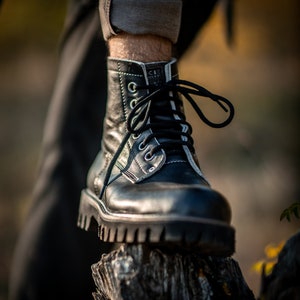 May include: Black leather boots with white stitching and black laces. The boots are worn by a person standing on a tree stump.