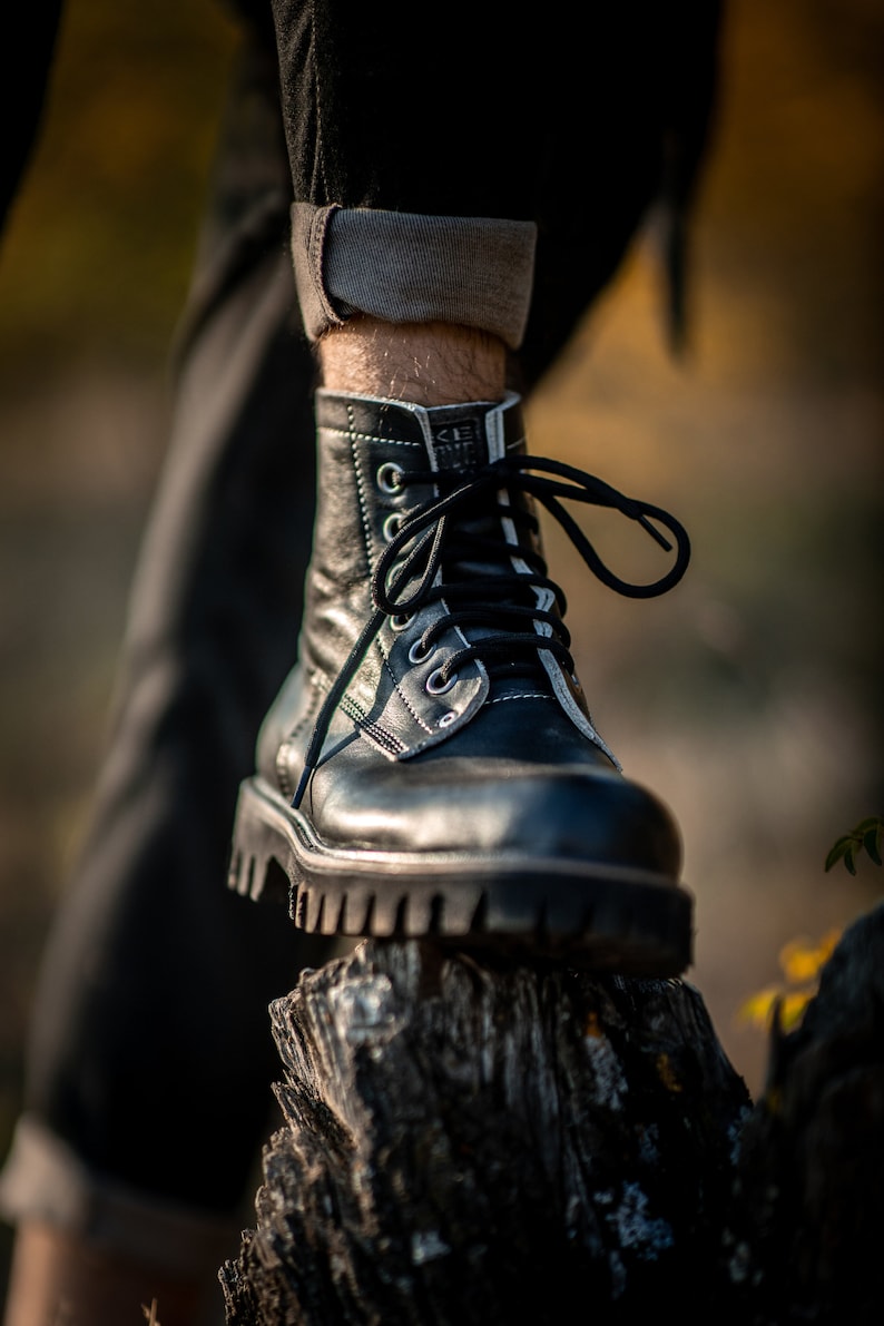 May include: Black leather boots with white stitching and black laces. The boots are worn by a person standing on a tree stump.