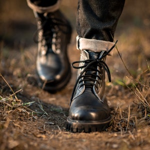 May include: Close-up of a pair of black leather boots with laces. The boots are worn by a person walking on a dirt path.