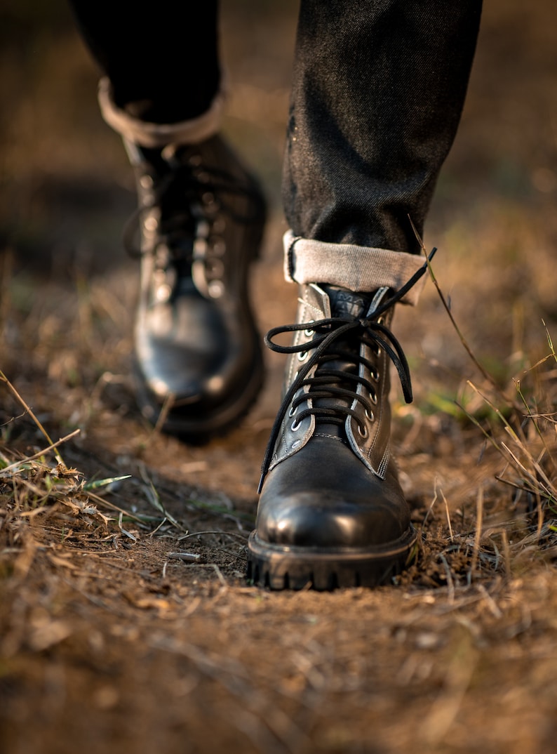 May include: Close-up of a pair of black leather boots with laces. The boots are worn by a person walking on a dirt path.