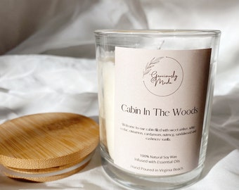 May include: A clear glass candle jar with a wooden lid. The candle is labeled "Cabin In The Woods" and has a scent description of "sweet amber, cedar, cinnamon, cardamom, nutmeg, sandalwood and cashmere vanilla". The candle is made with 100% natural soy wax and infused with essential oils. It is hand-poured in Virginia Beach.
