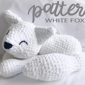 May include: A handmade white crochet fox toy with a smiling face and dark blue embroidered details. The text "pattern WHITE FOX" is in the upper right corner. The plush toy is lying down on a white surface.