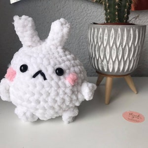 Op de afbeelding: Een handgemaakt wit gehaakt konijn met zwarte ogen, roze wangen en een kleine, zwarte, omgekeerde V-vormige mond. Het konijn heeft kleine armen en oren. Rechts staat een grijs en wit patroon pot met een cactus.