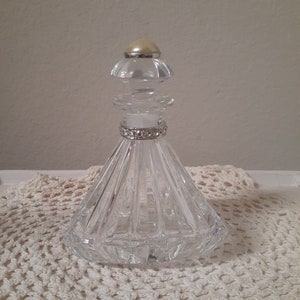 May include: A clear glass bottle with a diamond-shaped base and a crystal-like stopper. The bottle has a silver band with rhinestones around the neck.
