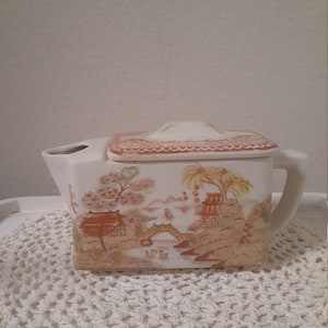 May include: Cream-colored ceramic teapot with a rectangular shape and a lid. The teapot features a scenic landscape design in shades of brown and tan, depicting a bridge, buildings, and trees. It has a handle and a spout.